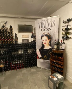 Amy Bailey Instagram - I was recently informed by a lovely fan that my enormous face from a poster signing at @germancomiccon now lives in their wine cellar.

I promise you, @nido1443 , that smug expression is one of camaraderie. She won’t judge. She may have killed others with a glass of wine or two, but you’re safe…as long as you remain a loyal subject, that is.
 ⚔️👸🏻💀

#DontDrinkTheWine