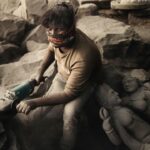 Arjun Kamath Instagram - The evening light was growing golden by the minute when I was driving through the quaint city of Khuldabad in Maharashtra. On the way, I saw an unusual whirlpool of dust outside my car window, so I decided to stop. As I cut through the flying dust, I saw a man engrossed in sculpting, his hands masterly guiding the machine that fervently chipped at a stone slab. Having acknowledged my presence, he carried on with the arduous task in all sincerity. I took the time to walk around and explore his petite workshop, which was decorated with half-made figurines and fine sculptures. The dusty haze of grey all around with the only colour being that of the man’s vibrant mask stood out for me, while the figurines in the background hinted at his work of the past few days. Intrigued by the whole setting, I quietly took the hold of my camera. In that exact moment, he looked up for the very first time and that was a sign enough for me to click on the shutter button of my camera. It was only later when we got talking that I could know more about him. His name was Hiralal Chauhan, and along with his family, he spent most of his time sculpting figurines out of kala patthar. Other talks were shared, about his work and how he has pursued it for 20 years. But the thing that stayed the most with me were his parting words. He told me that he was not able to pay due attention to me because his work demanded precise attention and a distraction of even a split second could result in grave injury. As he uttered those words, I was taken aback and could only see him in a light of greater respect. I believe that is what I love the most about travelling, that it allows me to learn more about the world and the people who live in it. Through my frames, I try to capture a sneak peek into people’s lives and tell their stories, capturing the essence of their personality. In a way, this act makes all my travels creatively fulfilling and fruitful, and I see it as one of the most enjoyable things about photography. #CapturedOnCanon with a Canon EOS 5D Mark IV & a 24-70mm F/2.8 II Lens #arjunkamathphotography #portrait #man #carving #stone #khultabad #aurangabad #yourshotphotographer Bhadra Maruti Temple, Khuldabad