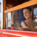 Özlem Yılmaz Instagram - Yaşım daha 16. Bakmayın öyle fotoğrafta takmış takıştırmış, o yaşta kokonalardan hallice göründüğüme. Daha çok underground partyler kovalıyormuşçasına giyindiğim bir tarzım vardı o günlerde😂 Bu fotoğraf sadece tek bir günün hikayesi, yıl 2002. Annem, arkadaşları, kuzenim.. Cümbür cemaat toplanıp Büyük Adaya gitmiştik. Yedik, içtik derken, kuzenimle kalktık masadan gezinmeye başladık. Az ilerde yüksek sesle konuşan biri, o zamanlar için adını bilmediğim malzemelerle yer değiştirip duran adamlar, patlayan flashlar, güzel kızlar.. Yaklaşıp aralarından birine “Ne yapıyorsunuz siz burada?” dedim Ali Kıran baş kesen gibi.. Ünlü bir altın markasının katalog çekimimiymiş? Dergi içinmiymiş neyse ne. “Hmm” dedim, döndük geri. Bir kaç adım sonra “Pardon” diye bir ses, bendeki de refleks arkamı dönüp bakmamla bir ışık patladı yüzümde. Fotoğrafım çekilmişti o an. Ardından bir kadın yanaşıp yarın yapacakları çekime katılıp katılamayacağımı sordu. Bende annemin yakında olduğunu, dilerse ondan izin istemesini söyledim. Geldi, izin istedi. Ayrıca karşılığında 200 Lira vereceklerdi. Kuzenimin yanımda gelmesi şartıyla annem ikna oldu ve biz, ertesi gün, kargalar henüz kahvaltılarını etmediği bir vakitte Taksimde olacağımız konusunda anlaştık. Gece oldu, gün oldu, yola çıktık. Toplanmalar, mekana geçmemiz, saç-makyaj derken çekimler başladı. Bir kaç kızdık. Kız derken ayıp olmasın, yanlarında kendimi mini mini minnoş hissettiğim ablalarlaydım 😂 Bir mekandan diğerine, ordan oraya sonunda ablaların çekimleri bitmiş, sıra bana gelmişti.. Durağan halde bir tramvaya oturttular beni. Sağa bak, sola bak, çekip duruyorlar poz poz. Çekim devam ederken, tesadüfün böylesi okuldan arkadaşım Feyza geçiyordu önümden. İster istemez baktı ne oluyor burada diye. O anda göz göze geldik. Saliselik affalasada, el kol hareketleriyle ve okuduğum ağzıyla orada ne yaptığımı sordu bir yandan yürümeye devam ederken. Çıkıp iki laflasam bişey derler miydi? Gülsem? İyiyim, anlar mı? Güldüm.. İşte bu kare öyle çıktı ortaya :) Ve gün bitti, artık eve gitme vakti.. Başta 200₺ dedikleri rakamı 100₺ olarak elime tutuşturdukları, geçmiş ve gelecek anılarıma bir yenisinide öylece eklediğim bir gün oldu benim için🙃