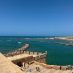 Vera Kolodzig Instagram – Saímos das ruínas de Volubilis já tarde e a chegada a Rabat foi já de noite. O grande desafio foi eu ver estrelinhas às cores a brilhar à minha frente por causa da minha falta de vista e lentes já cansadas. 
A capital de Marrocos é uma montra não muito representativa do resto do país e ainda assim encantadora! A vila nova distingue-se por avenidas de palmeiras alinhadas, a medina é de fácil circulação, a praia mesmo ali confere um aroma a maresia presente no ar, e o imponente Kasbah tem um ambiente sereno, tranquilo e limpinho (um kasbah é a cidadela das cidades árabes fortificadas). O turismo é pouco e maioritariamente local, mas na minha opinião vale a pena uma passagem por aqui para vaguear pelas avenidas largas e admirar as imponentes portas da cidade velha. Ainda nos atrevemos a um mergulho no mar, sendo claramente, as únicas duas europeias numa praia com centenas de pessoas. Para além disso ainda pudemos admirar o incrível trabalho de alguns artesãos que picam minuciosamente azulejos e criam os padrões tão tipicamente marroquinos.
Entretanto já nós fizemos à estrada outra vez, rumo a sul. Depois de uma paragem com temperaturas amenas, estamos preparadas para enfrentar o calor de Marraquexe.
.
.
.
#roadtrip #morocco #rabat #instatravel #travelwithfriends Rabat, Morocco