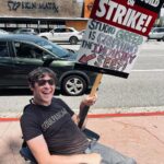 Zach Anner Instagram - As one of those writers and actors who works on several projects a year, but still can't make the minimum amount to get health insurance, this strike is pretty important to me! But it's not just about me obviously. It's about all the creative people who share their gifts and just want to make a living and be fairly compensated and appreciated for their work. #WGAStrong #sagaftra Image description: Zach, sitting in his wheelchair on a sidewalk in Burbank. he wears a shirt that reads "SOULPANCAKE" and hold a sign. At the top, it says "writers guild of America on strike" and at the bottom, in hand-drawn, colorful letters, it reads "studio greed, is crippling the industry! See?!" And then has an arrow pointing towards Zach in his wheelchair.