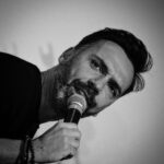 David Cristina Instagram - Parece que estava num comício do PSD, mas não. Estava a fazer um espectáculo numa festa de Natal de empresa. Isto de ser comediante tem muito de atuar para empresas porque o colégio dos meus filhos ainda não aceita integridade artística como pagamento. Há poucas coisas mais geradoras de ansiedade do que ser apresentado pelo CEO como uma “surpresa para os trabalhadores” e estar a caminhar para um palco, numa sala de conferências de hotel, rodeado de pessoas fortemente alcoolizadas que olham para nós com muita força para tentarem perceber de onde é que nos deviam conhecer. As festas de Natal de empresa são uma espécie de triângulo das Bermudas onde tudo pode acontecer. Um verdadeiro pesadelo para a Raquel dos Recursos Humanos que tem de fingir que o Vitor de Vendas (é sempre um Vitor de Vendas) não está a tentar comer a Carla da Contabilidade à frente de toda a gente. E a Carla, que é casada, mas num casamento infeliz com um senhor que passa mais tempo no campo de Padel do que a dar-lhe umas valentes raquetadas está a rir-se mais do que devia das anedotas que o Vitor sacou das Seleções do Reader’s Digest. Depois há o Zé que trata dos computadores da empresa há 30 anos e parou de estudar o assunto quando os ratos ainda tinham bolinha. Que acha que o Chat GPT é um carro desportivo e que decidiu que a cena dele esta noite ia ser pôr uma gravata na cabeça. Pelo meio temos a malta a recibos verdes que está a pôr comida nos bolsos e álcool no bucho porque as rendas em Lisboa estão como estão. E a servir esta maralha estão os empregados do hotel que têm toda a alegria de viver duma stripper de 65 anos que já viu tudo o que tinha a ver. Adoro festas de Natal de empresa, mas adoro ainda mais o maravilhoso constrangimento da segunda-feira seguinte. Isso sim, é uma “surpresa para os trabalhadores”.