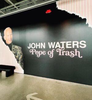 Seth Bogart Instagram - What an honor to design 3 collections of merchandise for the great new John Waters “Pope of Trash” show at @academymuseum ‼️
Seeing my art all huge on 2 walls at the end of the exhibit blew my mind. 

Love this pic @greggormanphoto took of John in front of my drawing of him. Just wait til you see the whole bathroom collection & the Divine/Pink Flamingos collection - both of those will be dropping sometime soon. Go see the show !!!