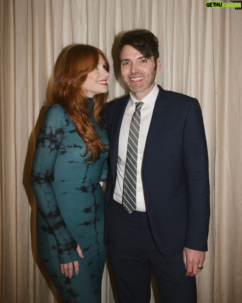 Bryce Dallas Howard Instagram - Today is 23 years since we started dating, and each day gets better and better. I love you handsome.⁣ ⁣ 📸: @andiejjane ⁣ [ID: Dressed up for a night out, BDH (left; wearing her hair down and a fitted dress with a moss & black dye print) and Seth Gabel (right; wearing a navy blue suit, white button-down shirt, and a gray and yellow tie) take a photo together in front of a tan curtain. BDH smiles softly at Seth.]