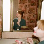 Bryce Dallas Howard Instagram - 💋💋💋⁣⁣⁣ ⁣ 📸: @andiejjane ⁣⁣⁣ [ID: In blue jeans and a teal linen blouse, BDH sits on her vanity by the mirror. She wears her hair up in a clipped bun and applies Bakeup Beauty’s neutral palm eyeshadow palette and Yolaine’s red lip.]