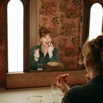 Bryce Dallas Howard Instagram - 💋💋💋⁣⁣⁣ ⁣ 📸: @andiejjane ⁣⁣⁣ [ID: In blue jeans and a teal linen blouse, BDH sits on her vanity by the mirror. She wears her hair up in a clipped bun and applies Bakeup Beauty’s neutral palm eyeshadow palette and Yolaine’s red lip.]