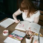 Bryce Dallas Howard Instagram - In Paradise 🌸 with my “Flowers, Feathers, & Animal Friends” ⁣ ⁣ Thank you Sarah (a.k.a @themintgardener) for yet another magical #WatercolorWorkbook – you make it so easy to put paint to paper and I can’t wait to fill every single page 💕⁣ ⁣ 📸: Andie Jane (@andiejjane)⁣ ⁣ [ID: Sitting at an outdoor patio table on a sunny day, BDH paints a pink and red rose in Sarah Simon’s new watercolor workbook, “Flowers, Feathers, & Animal Friends.” She wears a white t-shirt, glasses, and her hair down.]