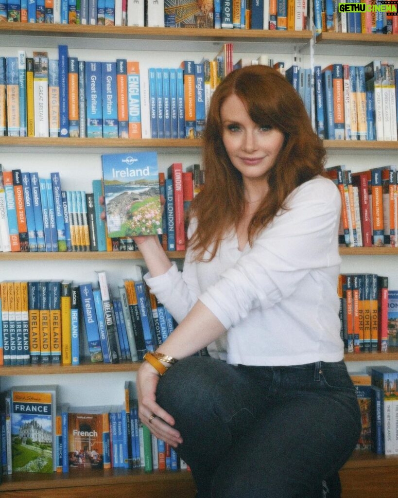 Bryce Dallas Howard Instagram - Returning to the motherland this week #Ireland 🇮🇪⁣⁣ ⁣⁣ 📸: @andiejjane⁣⁣ ⁣⁣ [ID: Wearing a white blouse and blue jeans, BDH stands in front of a bookcase with shelves and shelves of miscellaneous travel guides. She holds the Lonely Planet guide for Ireland by her face, absolutely ecstatic about returning to Ireland after 30 years!!]