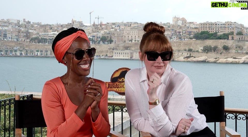 Bryce Dallas Howard Instagram - Going into the week like…⁣ ⁣ [ID: While in Malta for press, DeWanda Wise wears an apricot dress & BDH wears a white linen shirt. They make expressions that match their personalities to a tee: DeWanda smiles gleefully and Bryce leans toward her conspiratorially with a smirk. They sit in black directors chairs and behind them is the beautiful Maltese shoreline.]