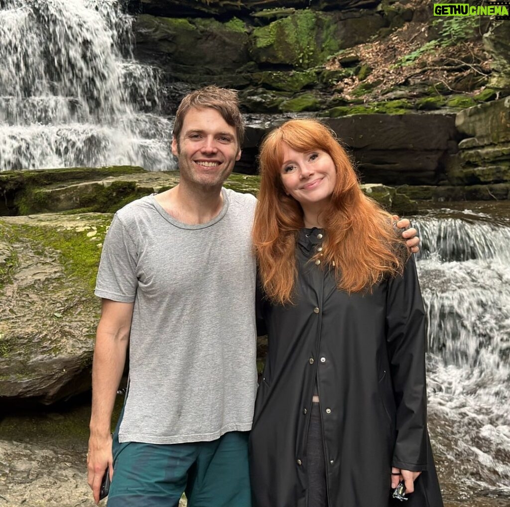 Bryce Dallas Howard Instagram - Happy Birthday handsome, I love you so much! @stealthgabel [ID: Standing in front of a rushing waterfall and mossy rocks in rural Pennsylvania, Seth Gabel (left) and BDH (right) wrap their arms around each other for a photo. Seth wears a grey t-shirt and teal swim trunks and BDH wears a black long raincoat and her hair down.]