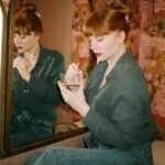 Bryce Dallas Howard Instagram - 💋💋💋⁣⁣⁣ ⁣ 📸: @andiejjane ⁣⁣⁣ [ID: In blue jeans and a teal linen blouse, BDH sits on her vanity by the mirror. She wears her hair up in a clipped bun and applies Bakeup Beauty’s neutral palm eyeshadow palette and Yolaine’s red lip.]