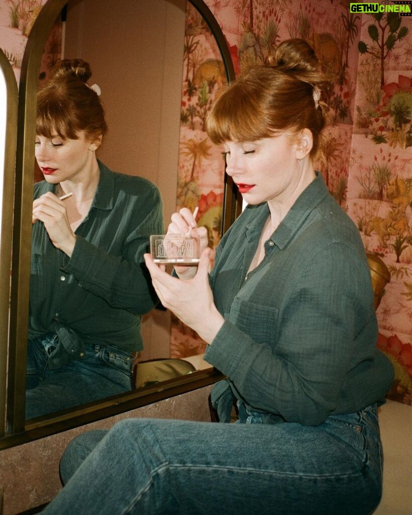 Bryce Dallas Howard Instagram - 💋💋💋⁣⁣⁣ ⁣ 📸: @andiejjane ⁣⁣⁣ [ID: In blue jeans and a teal linen blouse, BDH sits on her vanity by the mirror. She wears her hair up in a clipped bun and applies Bakeup Beauty’s neutral palm eyeshadow palette and Yolaine’s red lip.]