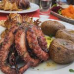 Laura Torrisi Instagram - Share roll (parte 2) Oltre a condividere altre foto del rullino, colgo l’occasione per parlarvi del cibo che ho trovato lungo quest’ avventura. Sapete che ho una passione per la cucina e se non fosse per la celiachia, assaggerei di tutto nei miei viaggi 😅 ma devo dire (così rispondo anche alle domande che mi hanno fatto alcuni di voi in merito) che Madeira è molto preparata sull’argomento! Con mio stupore, conoscono e hanno prestato molta attenzione anche alla “contaminazione” che solitamente durante i viaggi, mi trovo spesso a dover spiegare! 🙃 Cose da sapere se decidete di mangiare fuori: - L’alimentazione è prevalentemente dedicata alla carne e al pesce, alla griglia o preparati entrambi tipo zuppe o spezzatino, accompagnate spesso da ottime patatine fritte ma anche patate al cartoccio e purea di patate dolci o insalate miste e molto altro, tutti ottimi piatti e contorni con un unico ingrediente fisso: LA CIPOLLA 😂 quindi se non l’amate, fatelo presente al momento dell’ordinazione, perché la mettono davvero ovunque, cotta e sopratutto, cruda! Un piatto che sicuramente proverò a rifare a casa é “l’espetada” carne (di tutti i tipi) o pesce, cotta alla brace, messa a mo’ di spiedino su di uno stecco di alloro che conferisce alla pietanza un sapore davvero unico e particolare! A volte viene presentato al piatto a volte “appeso” al tavolo con degli appositi reggi spiedini di metallo molto scenografici. Ottime ed invitanti opzioni anche per vegani e segnalo anche una varietà di frutti tropicali da fare invidia! 🥑 Unica pecca: il prezzo! Veramente folle!!! 🫠 A mio avviso, é più convenite mangiare fuori rispetto a fare la spesa perché i prezzi sono molto simili a quelli che abbiamo in Italia, mentre mangiando fuori (bene) spenderete 30/35€ a coppia circa. Comunque vale sempre la pena assaggiare i piatti tipici del posto 😊 Spero di esservi stata utile! 🤗 Ps: per i celiaci e/o gli intolleranti al glutine: a Funchal c’è un negozio interamente senza glutine dove trovate, dolce e salato (confezionato) e qualcosa di fresco ma surgelato! Io ho preso dell’ottimo pane in cassetta che mi ha fatto stracomodo per i panini spezza trekking! 😎
