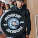 Tia-Clair Toomey Instagram – Sunday, 1:34pm: The fact that there is now a whole competition floor on the Dell Diamond concourse where a concession stand used to be — complete with 5 sets of double-under and lifting platforms, barricades and a sharp-looking @roguefitness backdrop — feels so natural, it’s easy to assume this was always the plan. It was not, but after four days of Book of Job-like weather, @caityhenniger and @billhenniger are taking no chances for the final event. 

Moving “The Cleanup” from the field to the concourse at the last minute is the latest example of the kind of logistical sorcery the @RogueInvitational has been performing behind the scenes all weekend. But not even Rogue can change the temperature, which dropped from 75° to 50° in the span of an hour earlier this morning. The top heat of women, the last of four heats, spend most of their 45 minute wait trying to stay warm; when @alexgazan_ 🇺🇸 takes a chair into the women’s bathroom, @laurahorvaht 🇭🇺 and @gabimiga 🇵🇱 follow suit. 

@tiaclair1 🇦🇺 remains outside, pacing around the warmup area with @shaneorr01’s jacket draped across her shoulders like a cape, superhero style. Shane, down to a just a longsleeve t-shirt, paces with her, shivering so hard he’s practically vibrating. A member of the Rogue event team notices, removes his own brand new Invitational hoodie and offers it to Shane, who accepts it with a mix of surprise and gratitude. By the time he pulls it on, @nick.p.jeffries is back at work, the only person on the concourse wearing shorts and a t-shirt.

If you want to know what people mean when they say the Rogue Invitational goes above and beyond — it’s this.

✍🏽: @christinedca
📷: @emomphoto